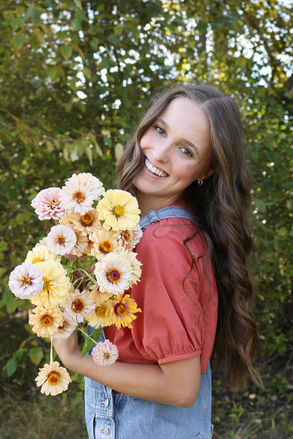 A young woman holding a bouquet of specialty zinnia flowers in front of a green leafy background with a pink blouse on and overalls. She is smiling wile holding flowers