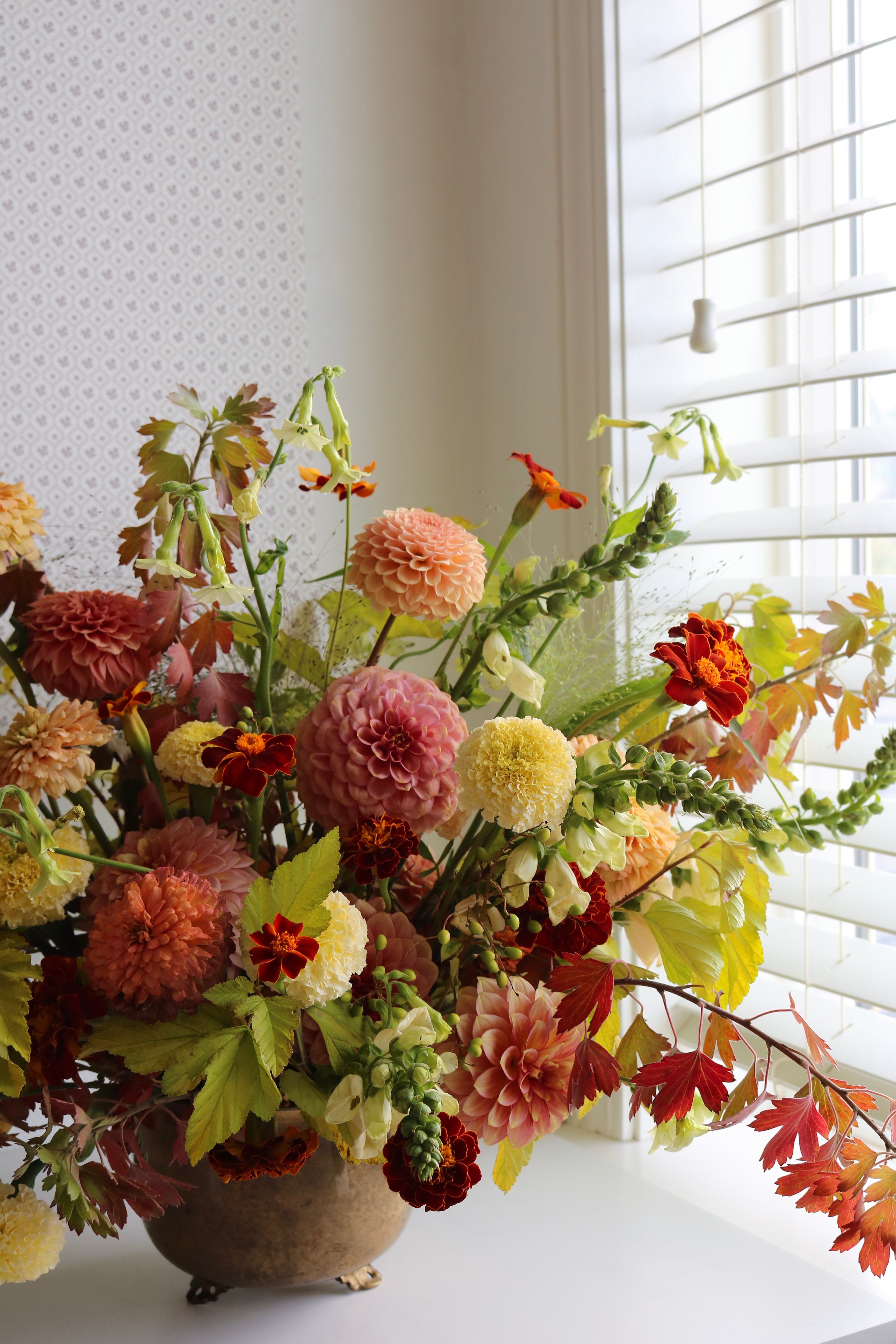 An autumnal floral arrangement in a rustic brass footed bowl, with shades of orange, red, pink, yellow, and green, displayed on a windowsill with white blinds. This inlcudes River Merle dahlias, foxglove, ninebark branches, marigolds, and zinnias.